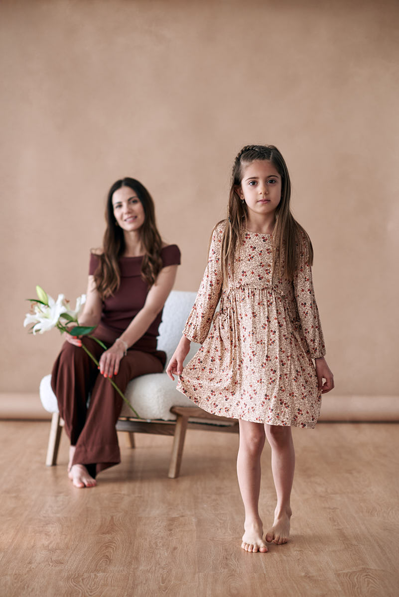 Retrato de madre e hija en un entorno cálido Madre sentada en un sillón, sosteniendo flores, mientras su hija pequeña se para frente a ella, todo en un fondo cálido y delicado, ideal para una sesión de fotos del Día de la Madre.