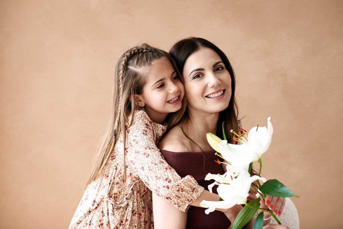 Sesión fotográfica de madre e hija con flores Madre e hija sonriendo, la madre sostiene un ramo de lirios mientras su hija abraza su espalda en un ambiente cálido y acogedor para una sesión de fotos del Día de la Madre.