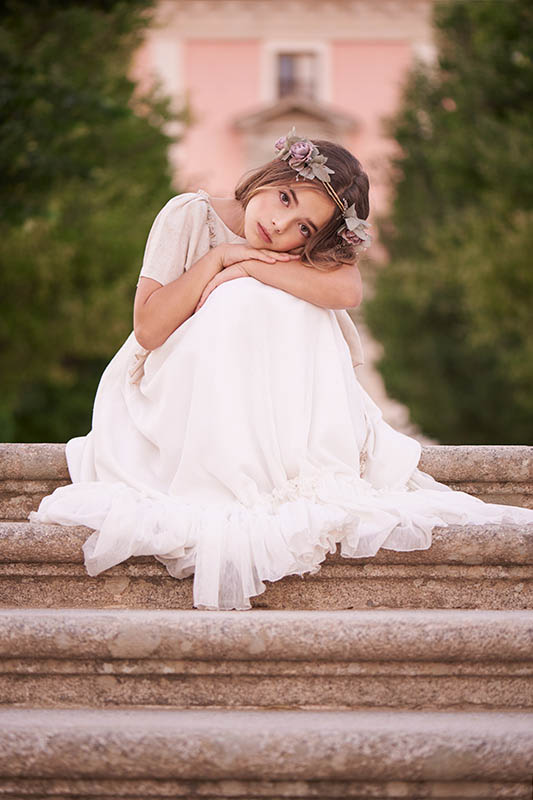 Niña con vestido de comunión blanco sentada en escaleras al aire libre durante sesión en Madrid.