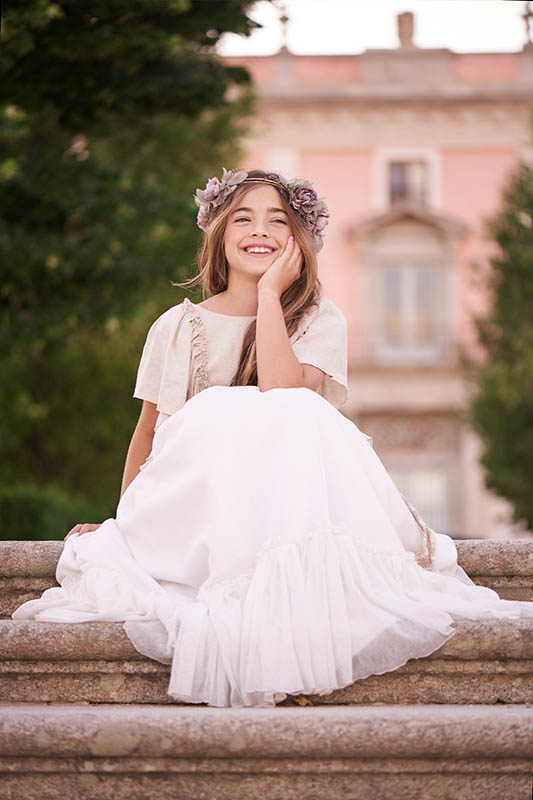 Niña con vestido blanco de comunión sentada en el suelo frente a un edificio clásico en Madrid.