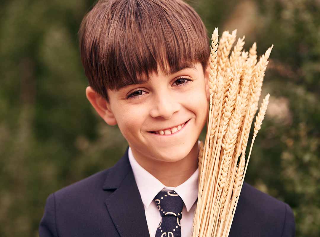 Niño vestido de comunión con traje negro sosteniendo espigas en un jardín en Madrid.