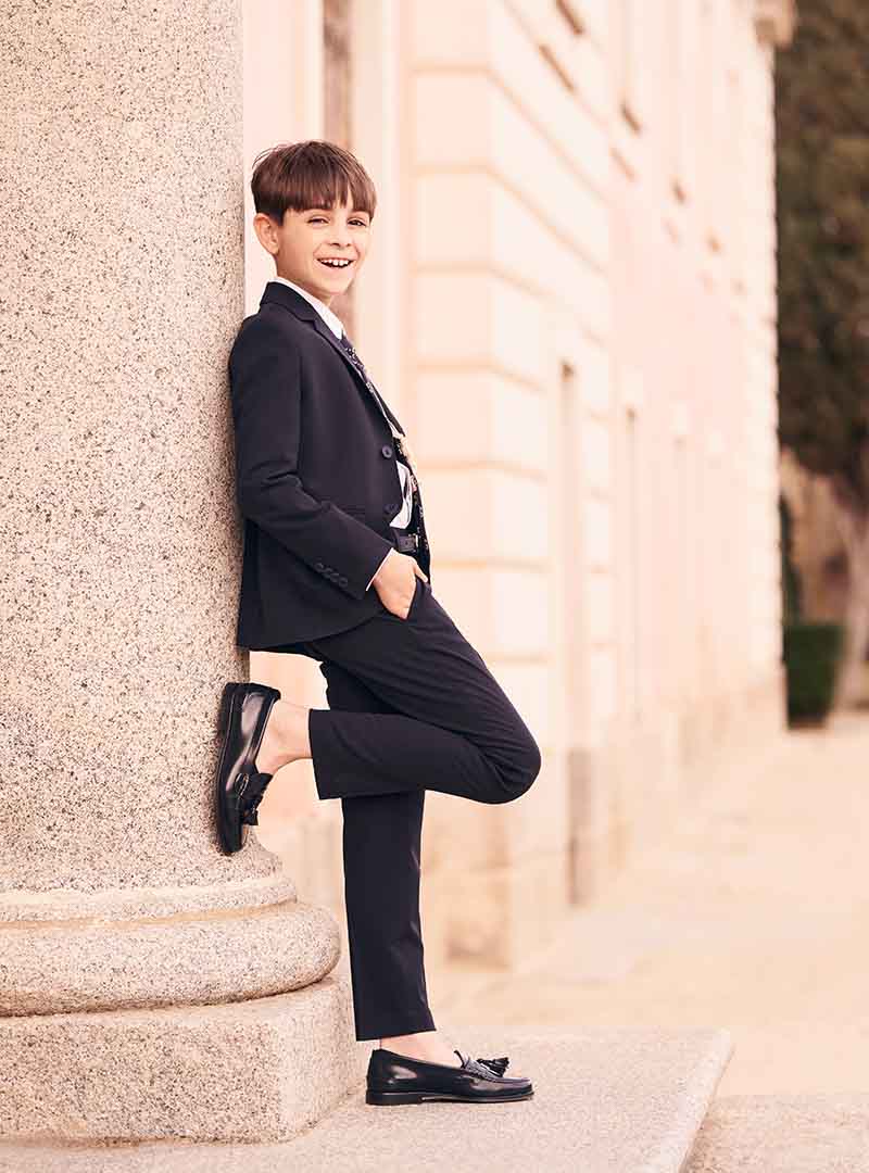 Niño con traje negro de comunión posando apoyado en una pared al aire libre en Madrid.