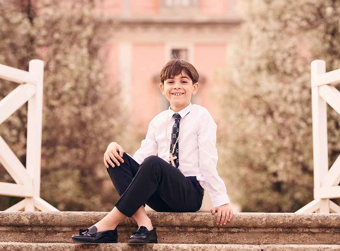Niño vestido de comunión sonriendo junto a una barandilla con un fondo clásico en Madrid.
