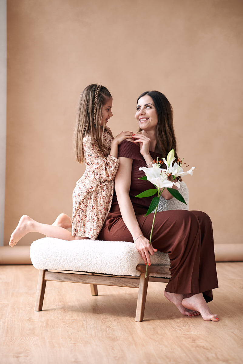 Retrato familiar de madre e hija en sesión fotográfica Madre sentada en un sillón mientras su hija pequeña la abraza por detrás, en una sesión de fotos del Día de la Madre.