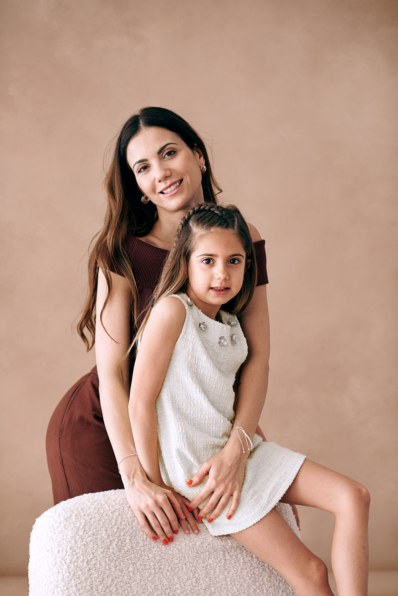 Madre e hija en pose tierna para el Día de la Madre Madre e hija sonrientes, sentadas en un sillón, mostrando una conexión cercana mientras se abrazan, con flores en la mano, capturando la esencia de una sesión de fotos para el Día de la Madre.